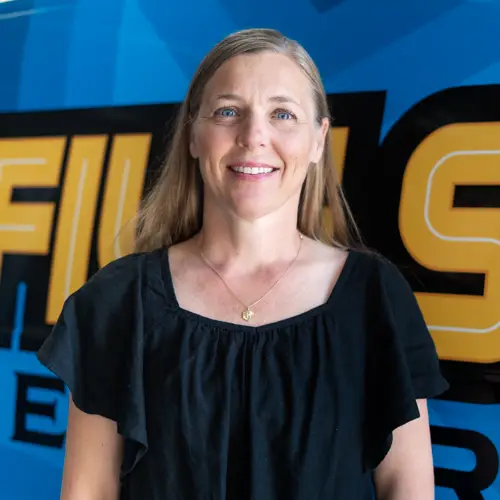 Kristen webp A woman with long hair is standing in front of a blue and yellow wall featuring bold lettering, wearing a black top with ruffled sleeves.